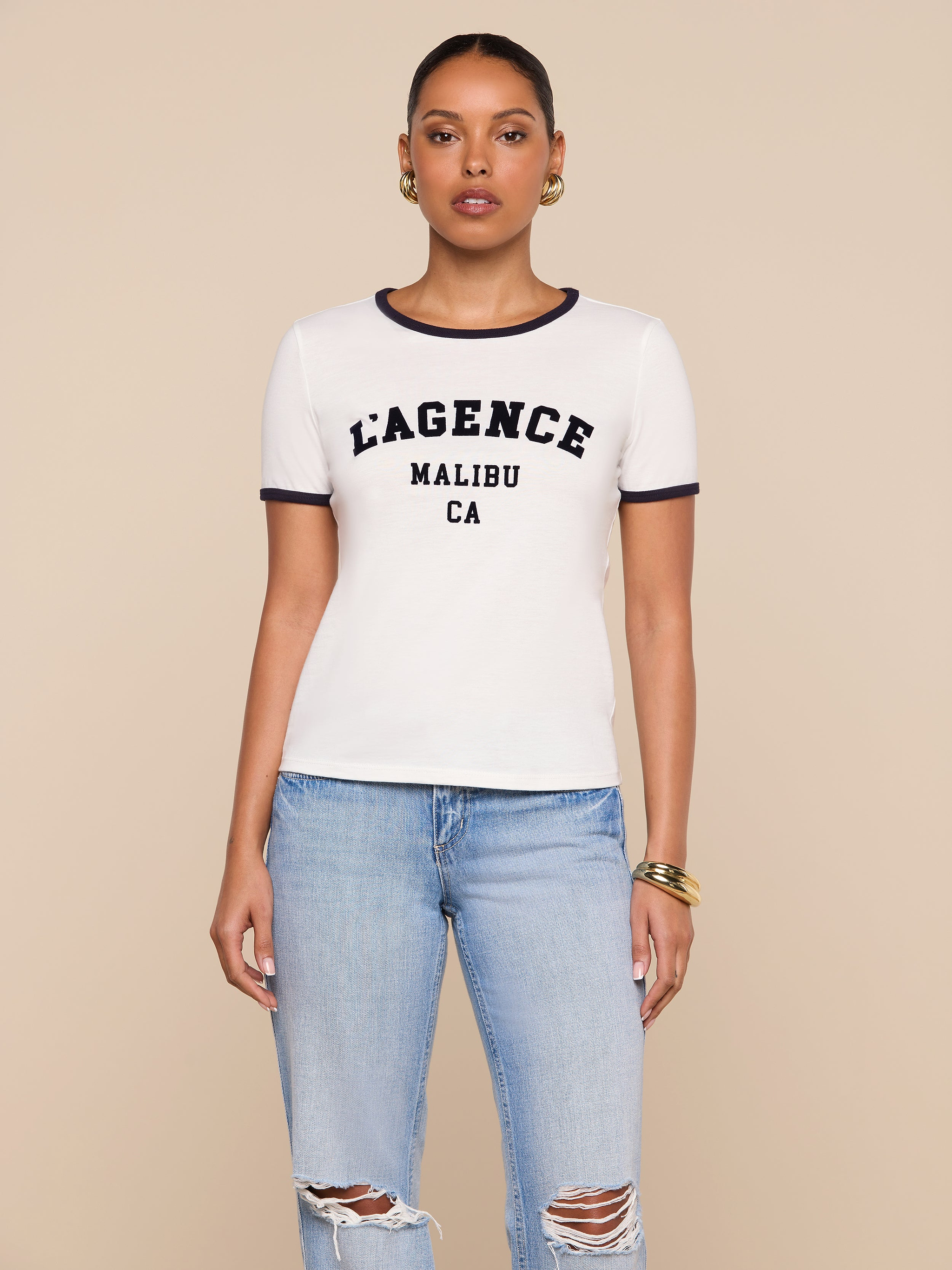 A woman stands against a beige background wearing a white Tommie Crewneck Tee featuring bold L’AGENCE MALIBU CA lettering, light blue ripped jeans, gold hoop earrings, and gold bangles.