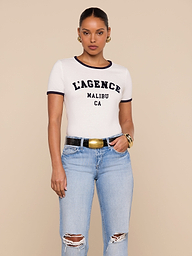 A woman stands against a beige background, wearing a white Tommie Crewneck Tee with LAGENCE MALIBU CA printed on it, light blue ripped jeans, a black belt with a gold buckle, gold hoop earrings, and a gold bracelet.