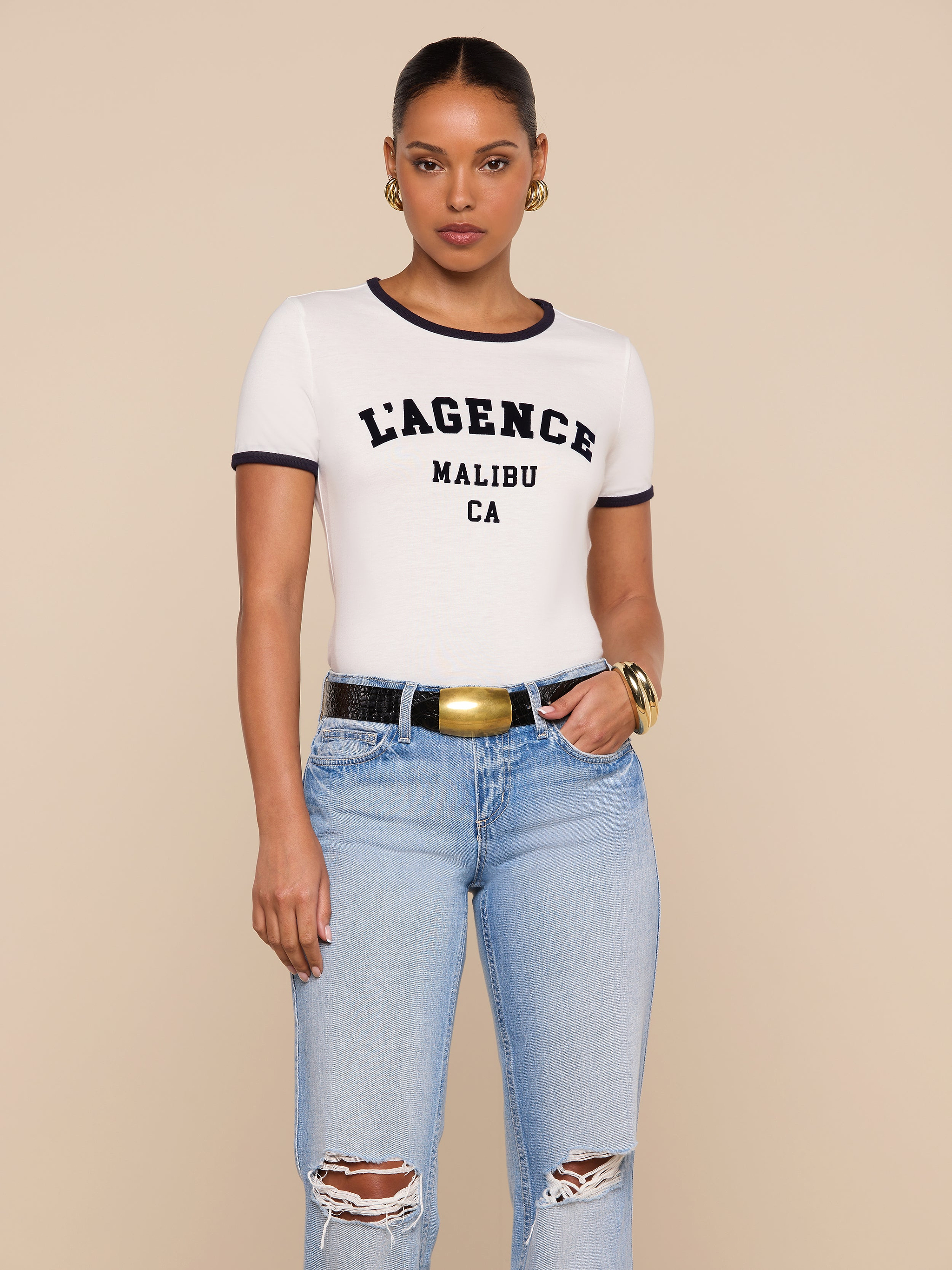 A woman stands against a beige background, wearing a white Tommie Crewneck Tee with LAGENCE MALIBU CA printed on it, light blue ripped jeans, a black belt with a gold buckle, gold hoop earrings, and a gold bracelet.