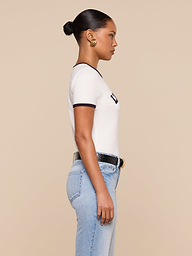 A woman stands in profile against a beige background, wearing a white Tommie Crewneck Tee with black trim, light blue high-waisted jeans, a black belt with a gold buckle, gold earrings, and her hair styled in a sleek bun.