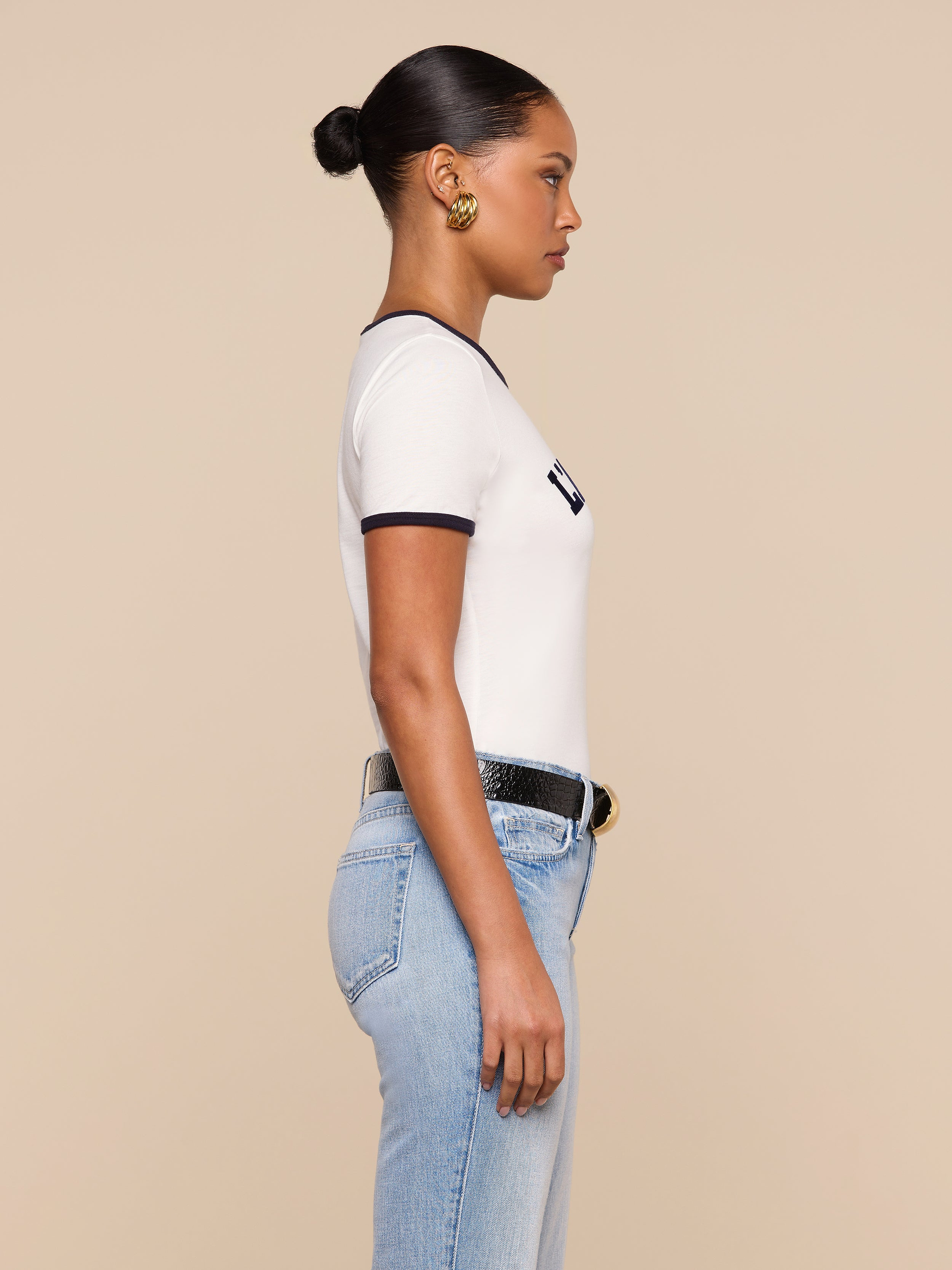 A woman stands in profile against a beige background, wearing a white Tommie Crewneck Tee with black trim, light blue high-waisted jeans, a black belt with a gold buckle, gold earrings, and her hair styled in a sleek bun.