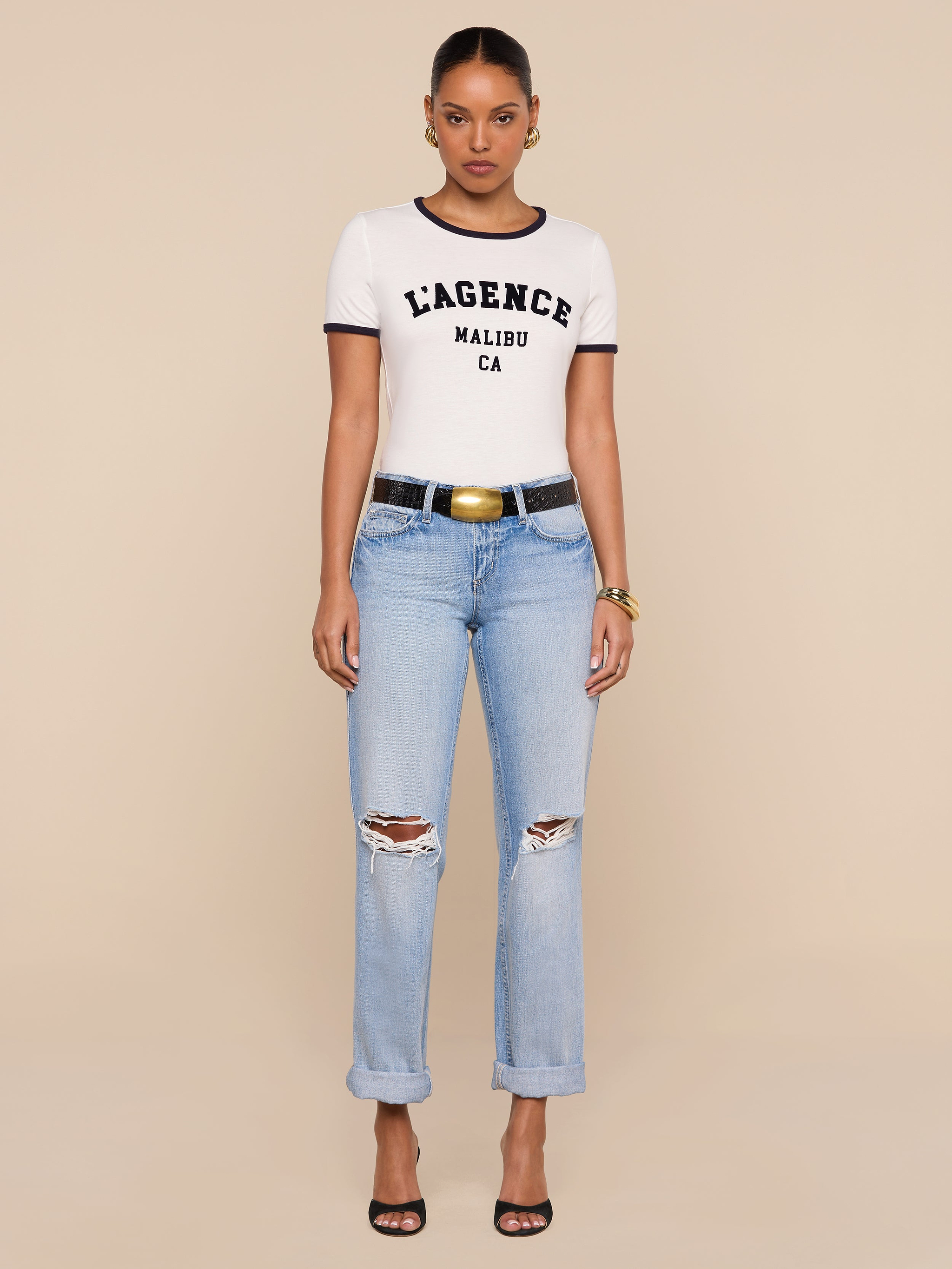 A woman stands against a beige background wearing a white Tommie Crewneck Tee, light blue ripped jeans with rolled cuffs, a black belt with a gold buckle, hoop earrings, gold bracelets, and black heels.