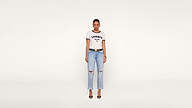 A woman stands facing forward, wearing a white KASENCE MALIBU CA t-shirt, light blue ripped jeans, black belt, and black heels against a plain white background.