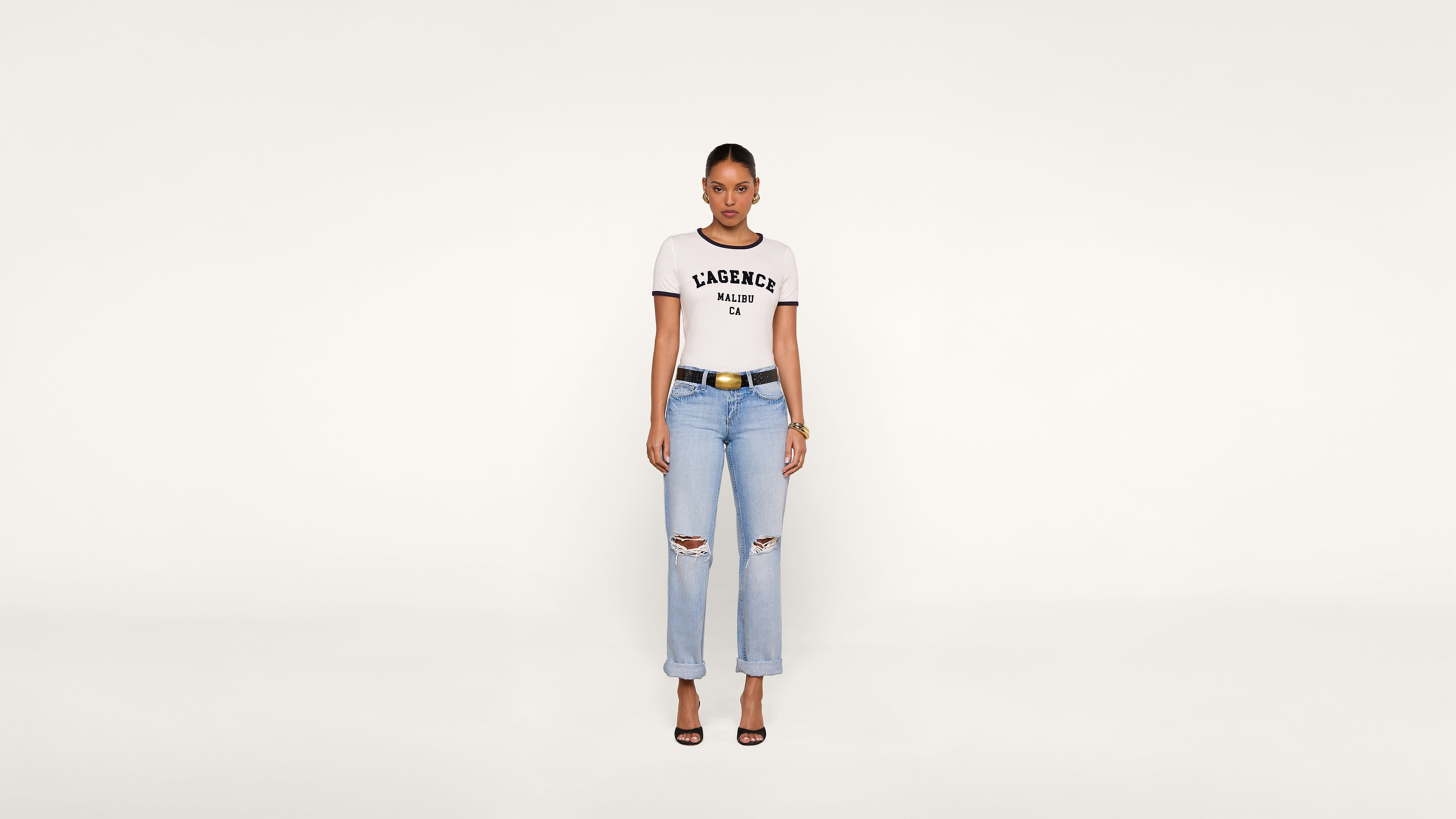 A woman stands facing forward, wearing a white KASENCE MALIBU CA t-shirt, light blue ripped jeans, black belt, and black heels against a plain white background.