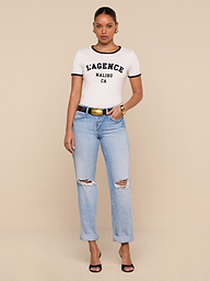 A woman stands against a beige background wearing a white Tommie Crewneck Tee, light blue ripped jeans with rolled cuffs, a black belt with a gold buckle, black heels, and gold hoop earrings and bracelets.