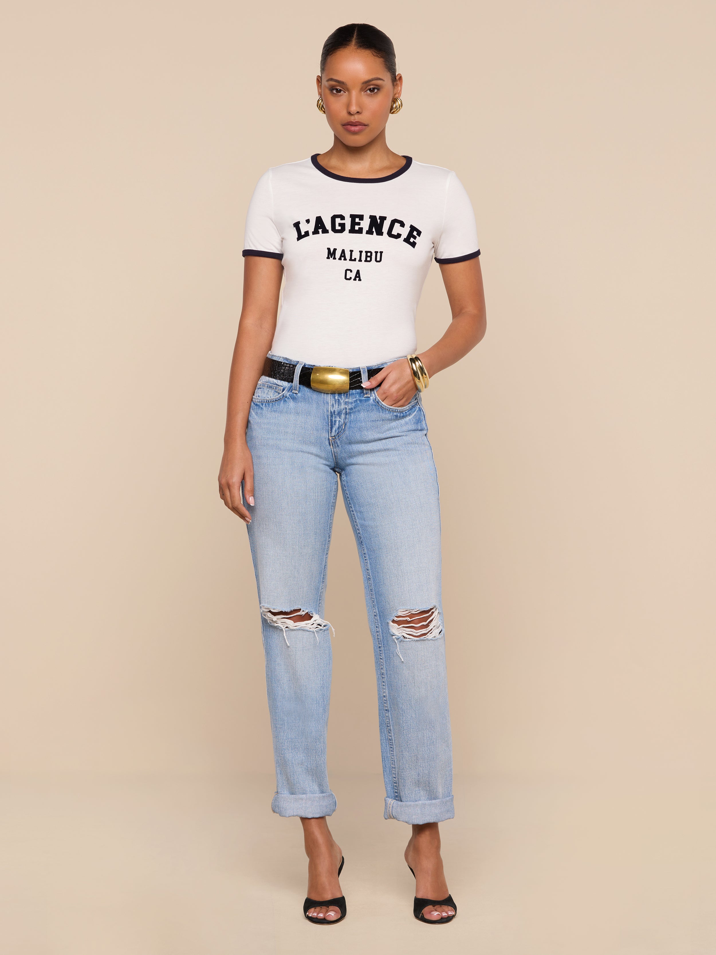 A woman stands against a beige background wearing a white Tommie Crewneck Tee, light blue ripped jeans with rolled cuffs, a black belt with a gold buckle, black heels, and gold hoop earrings and bracelets.