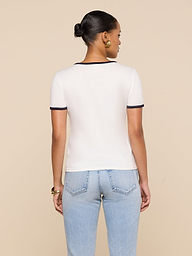 A woman with her hair in a bun stands with her back to the camera, wearing a white Tommie Crewneck Tee with dark trim and light blue jeans against a beige background.