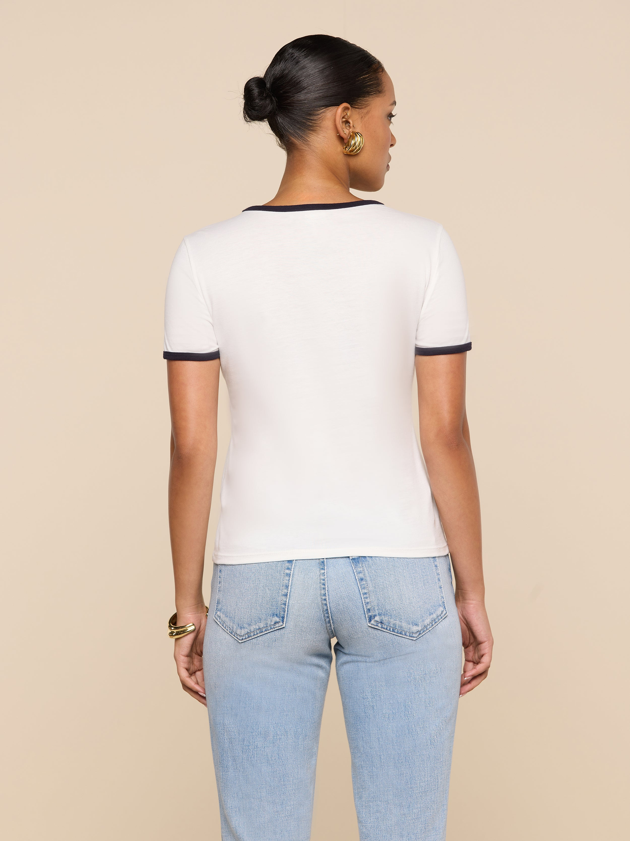 A woman with her hair in a bun stands with her back to the camera, wearing a white Tommie Crewneck Tee with dark trim and light blue jeans against a beige background.