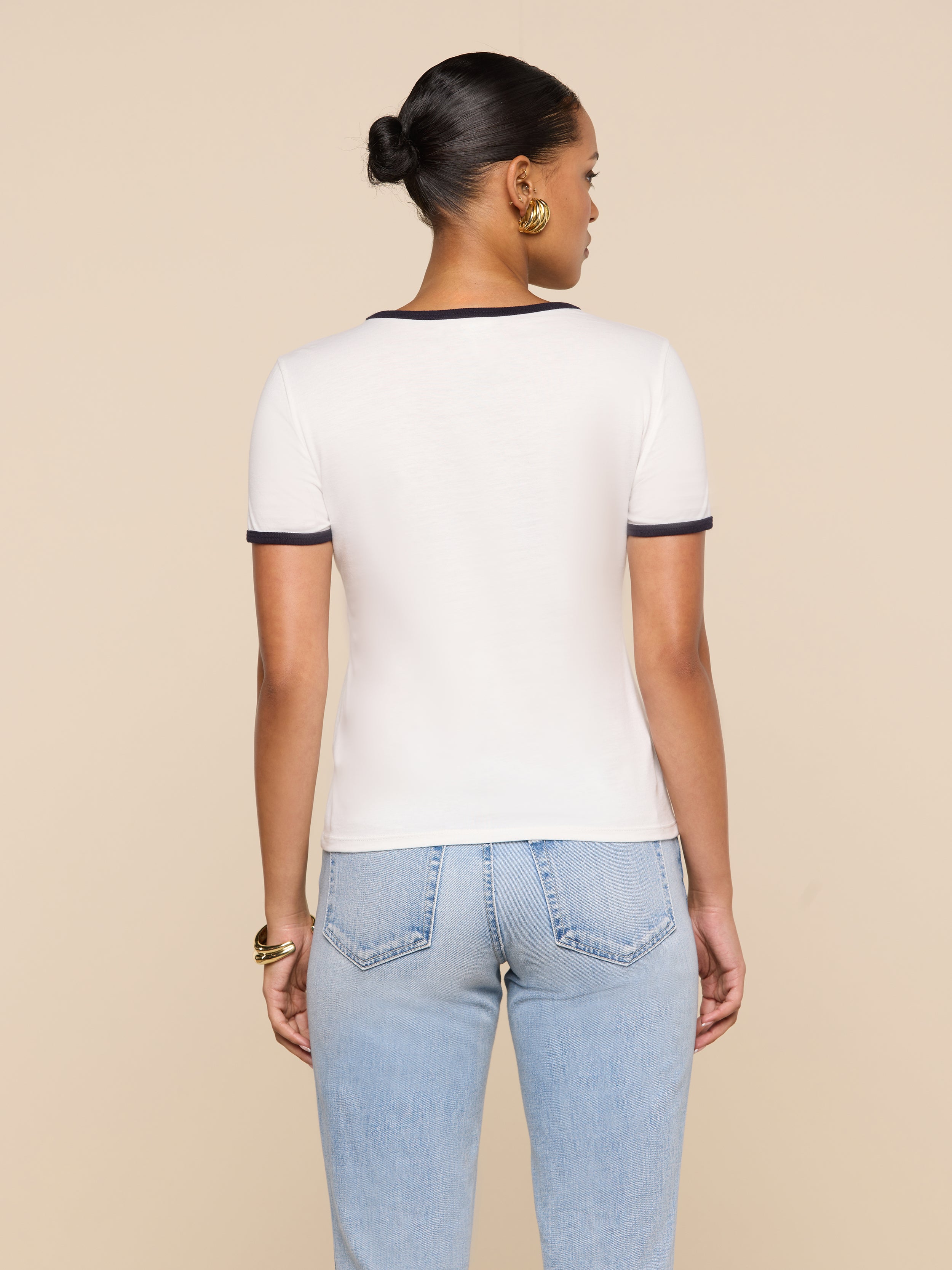 A woman with her hair in a bun stands with her back to the camera, wearing a white Tommie Crewneck Tee with dark trim and light blue jeans against a beige background.