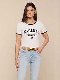 A woman wearing the Tommie Crewneck Tee—a relaxed fit white ringer tee made from Supima cotton featuring LAGENCE NEWPORT CA—paired with light blue jeans, a tan belt with a gold buckle, and gold hoop earrings, stands against a plain background.