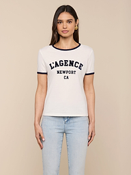 A woman is wearing the Tommie Crewneck Tee—a white short-sleeve ringer shirt with black trim and “LAGENCE NEWPORT CA” printed on the front, crafted from soft Supima cotton—paired with light blue jeans and standing against a plain white background.