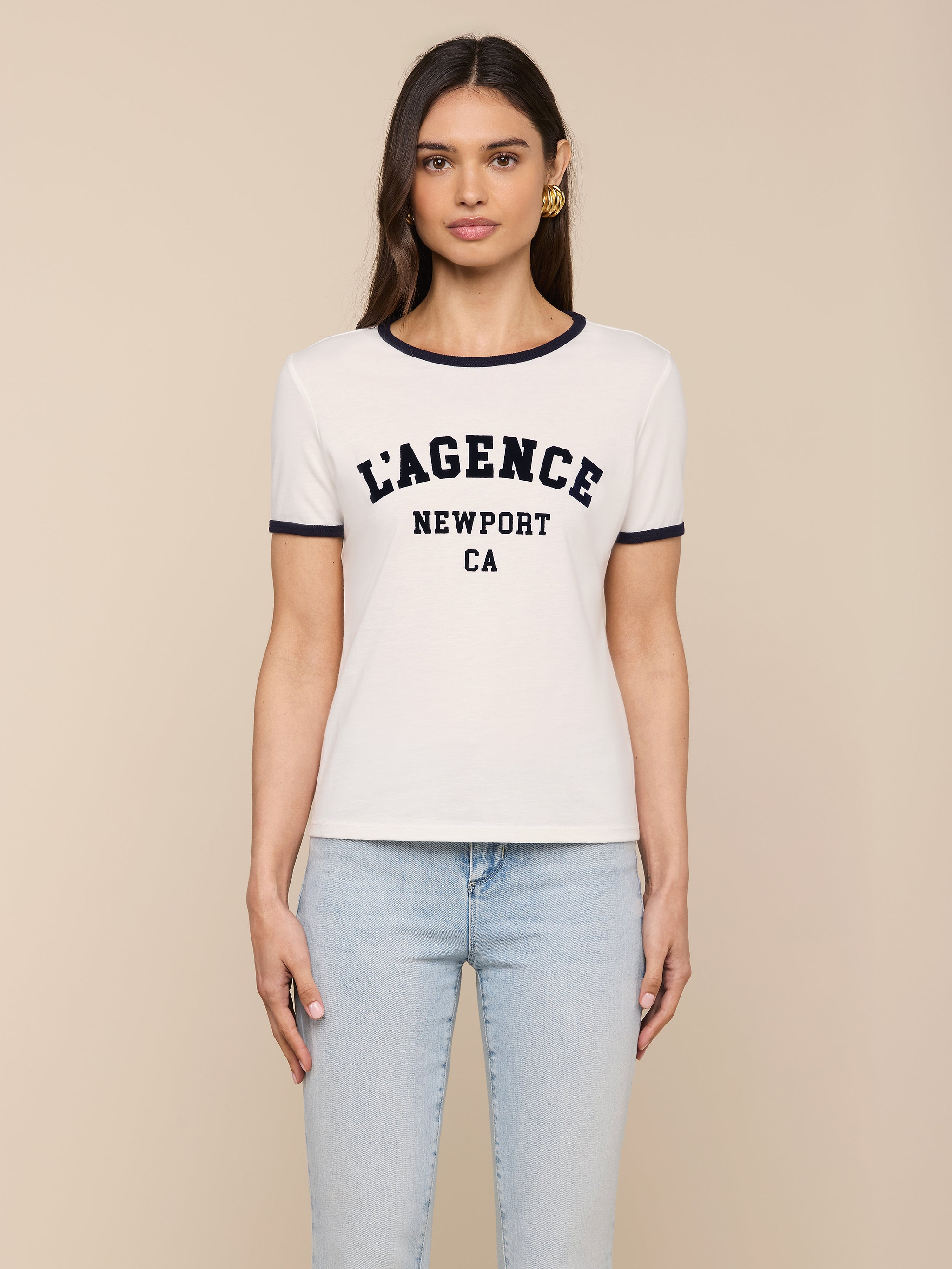 A woman is wearing the Tommie Crewneck Tee—a white short-sleeve ringer shirt with black trim and “LAGENCE NEWPORT CA” printed on the front, crafted from soft Supima cotton—paired with light blue jeans and standing against a plain white background.