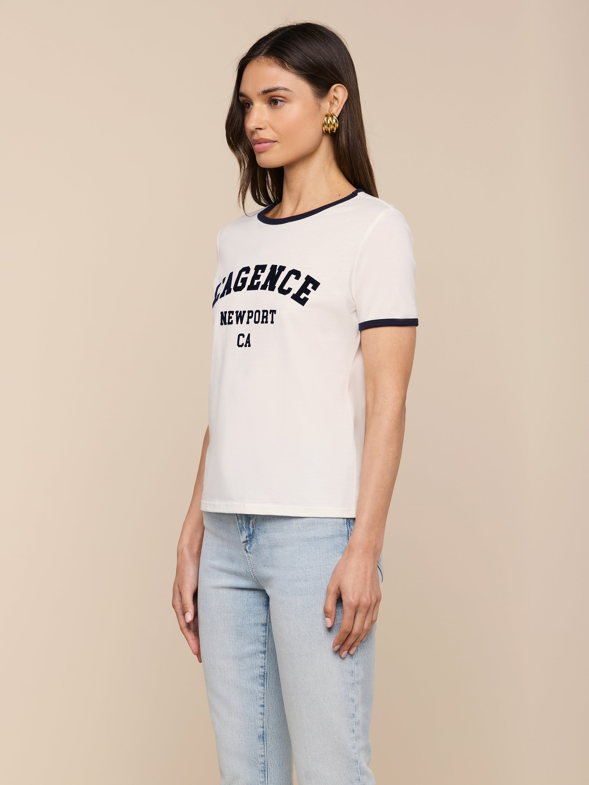 A woman wearing the Tommie Crewneck Tee, a white ringer t-shirt with “LAGENCE NEWPORT CA” text, paired with relaxed fit light blue jeans and gold earrings, stands against a plain light background facing slightly to the side.