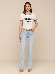 A woman stands against a white background wearing a Tommie Crewneck Tee, light blue jeans, a tan belt, and tan heels. She faces forward with arms relaxed at her sides in a relaxed fit ensemble.