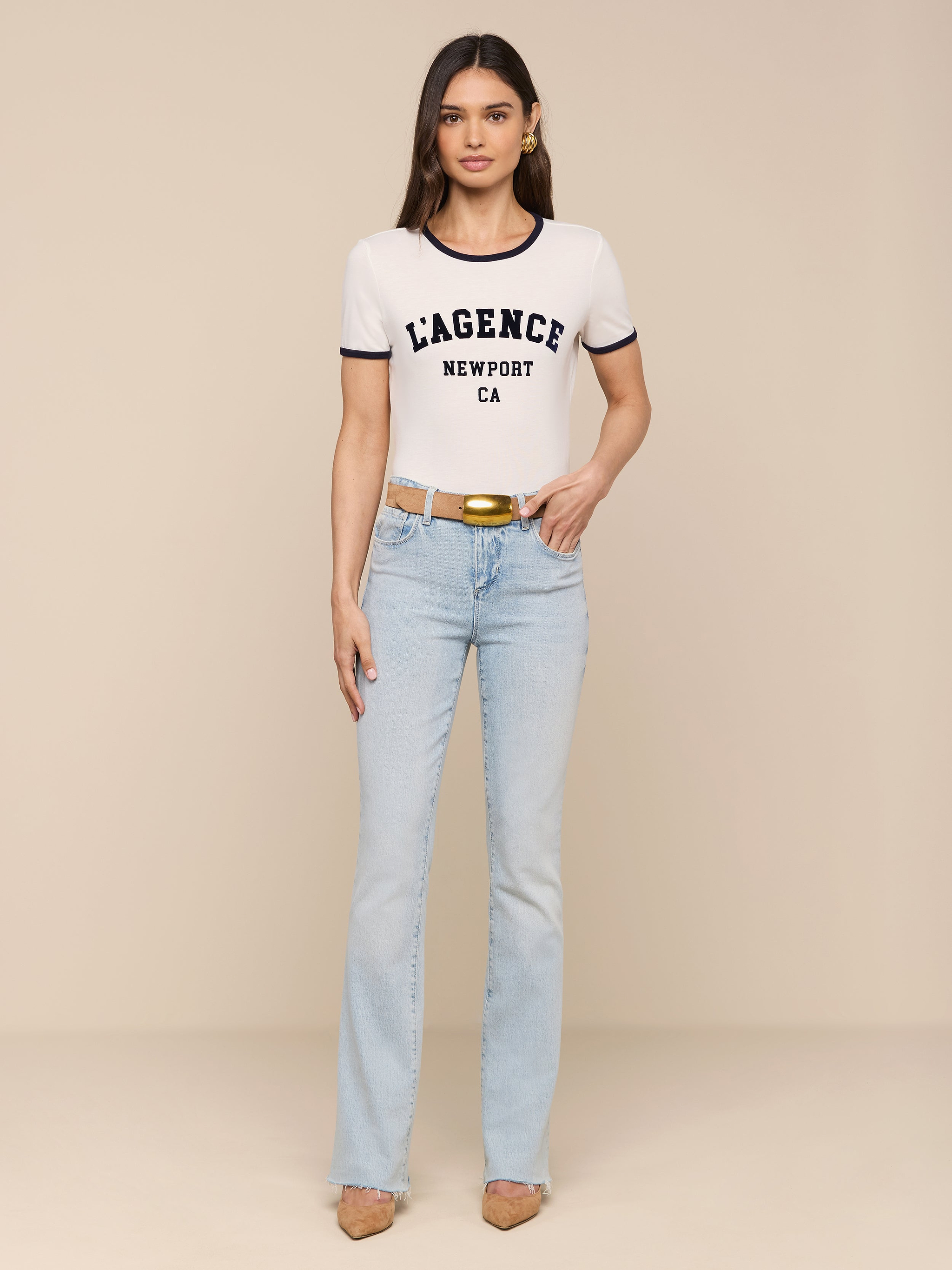 A woman stands against a white background wearing a Tommie Crewneck Tee, light blue jeans, a tan belt, and tan heels. She faces forward with arms relaxed at her sides in a relaxed fit ensemble.