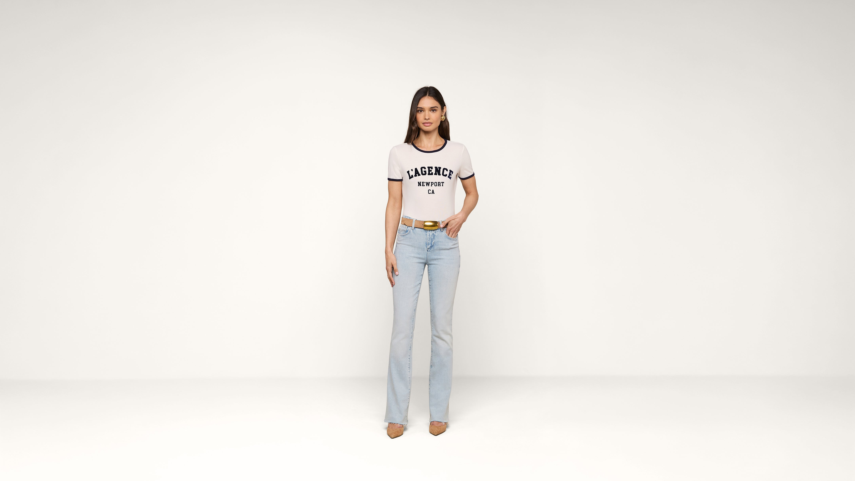A woman stands in a plain white room, wearing a white graphic t-shirt, light blue jeans, and beige shoes. She has straight brown hair and a relaxed expression, with one hand on her hip and the other by her side.