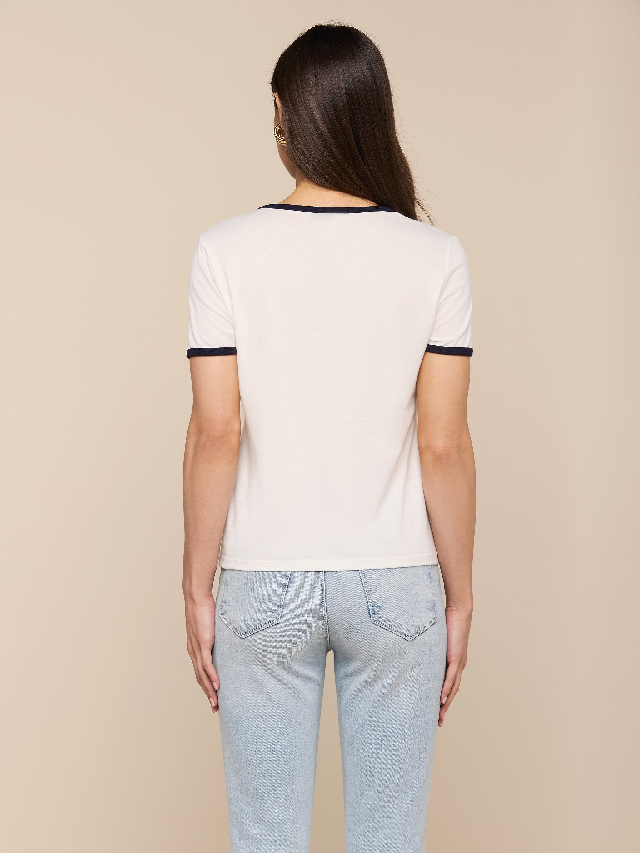 A person with long brown hair, wearing the Tommie Crewneck Tee— a relaxed fit white ringer tee with dark trim—paired with light blue jeans is standing with their back to the camera against a plain background.