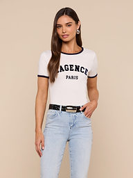 A woman with long brown hair wears the Tommie Crewneck Tee— a white, short-sleeve Paris top made from soft Supima cotton that says AGENCE PARIS—paired with light blue jeans, a black belt, and hoop earrings, standing against a plain light background.