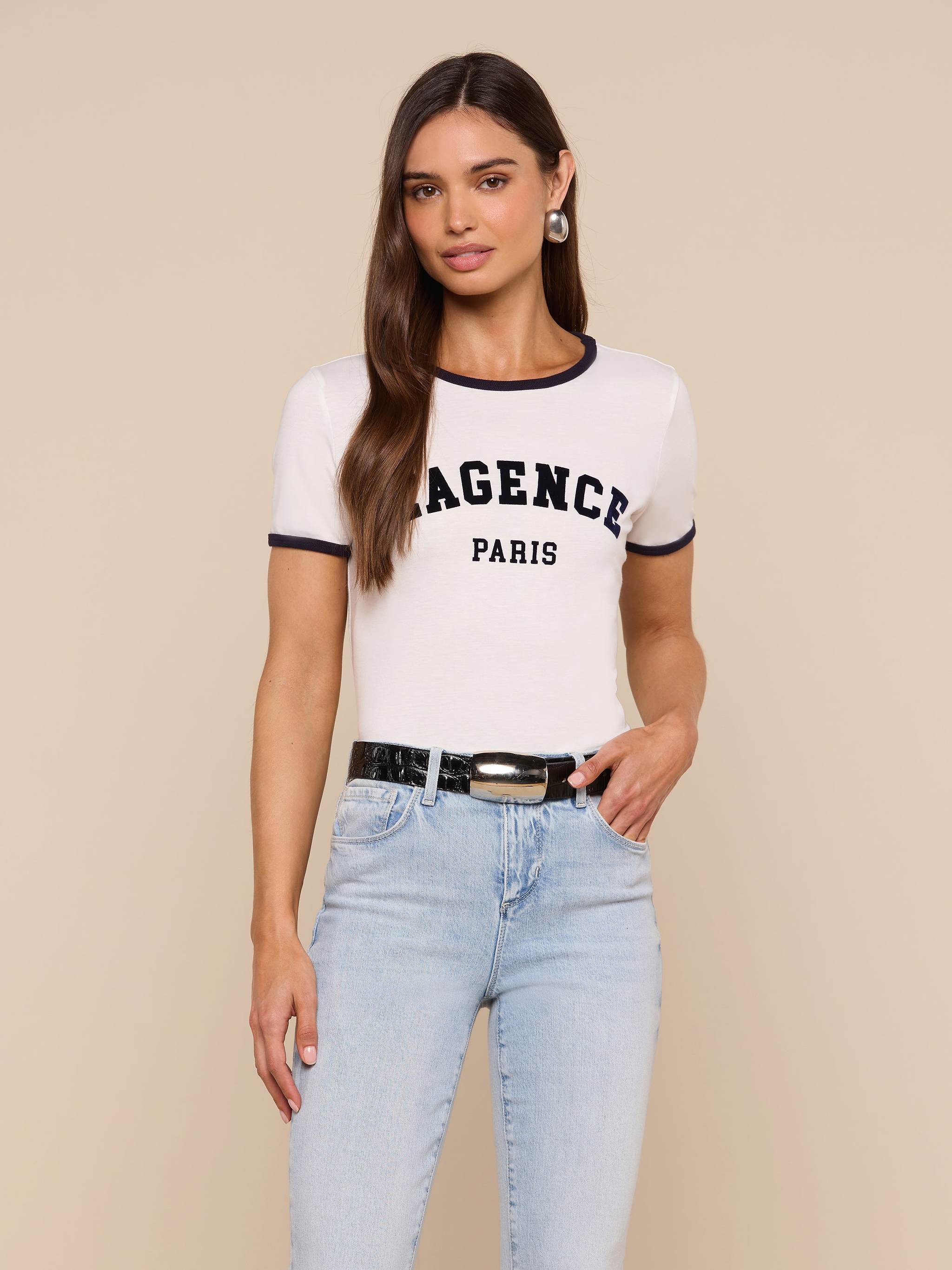 A woman with long brown hair wears the Tommie Crewneck Tee— a white, short-sleeve Paris top made from soft Supima cotton that says AGENCE PARIS—paired with light blue jeans, a black belt, and hoop earrings, standing against a plain light background.