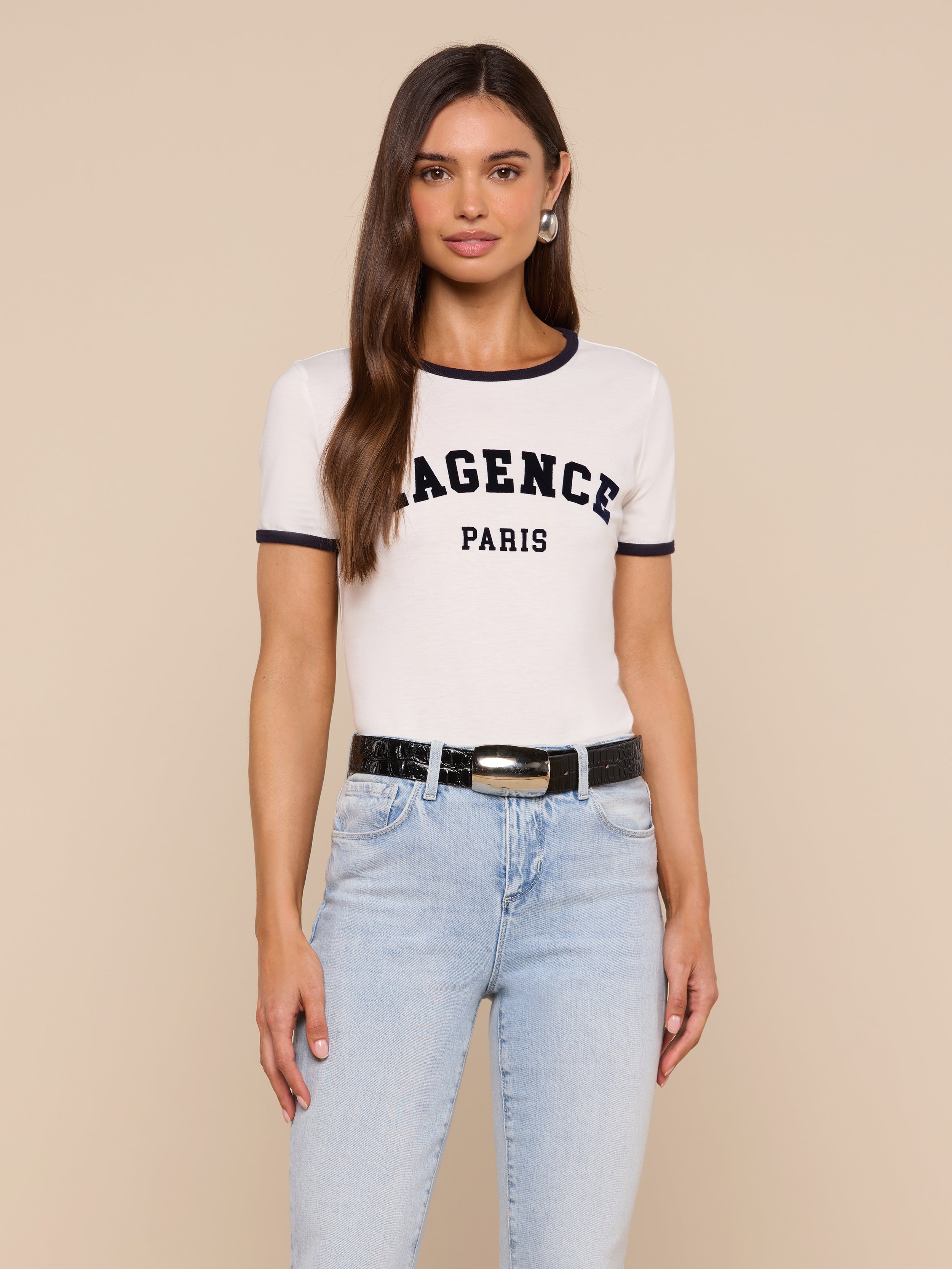 A woman with long brown hair wears a white Tommie Crewneck Tee crafted from Supima cotton, styled with light blue jeans and a black belt, standing against a plain light background.