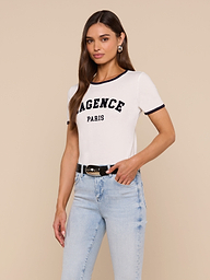 Woman wearing the Tommie Crewneck Tee in white with LAGENCE PARIS text, light blue jeans, black belt, and hoop earrings, standing against a plain light background in a relaxed fit Supima cotton style.