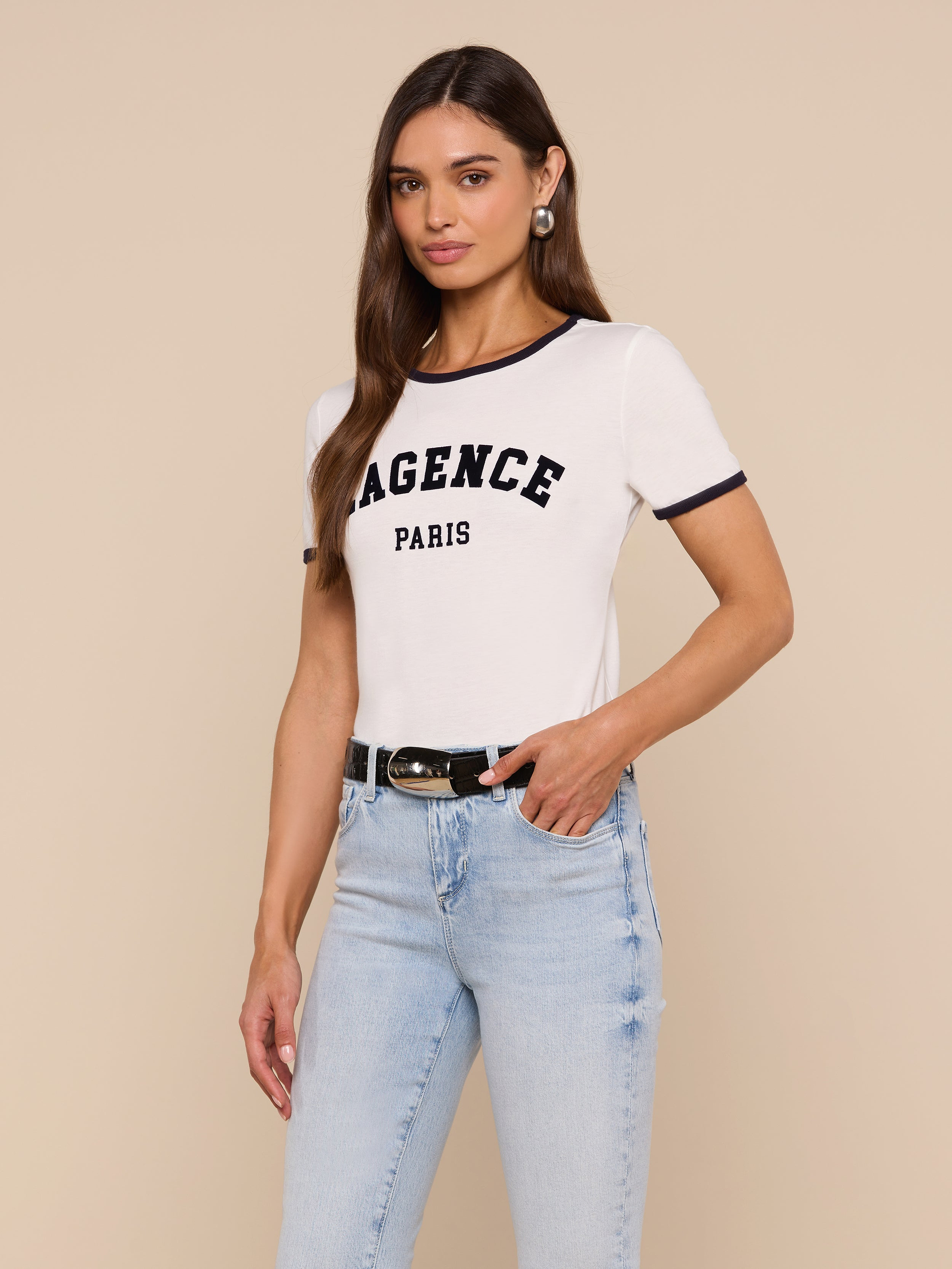 Woman wearing the Tommie Crewneck Tee in white with LAGENCE PARIS text, light blue jeans, black belt, and hoop earrings, standing against a plain light background in a relaxed fit Supima cotton style.