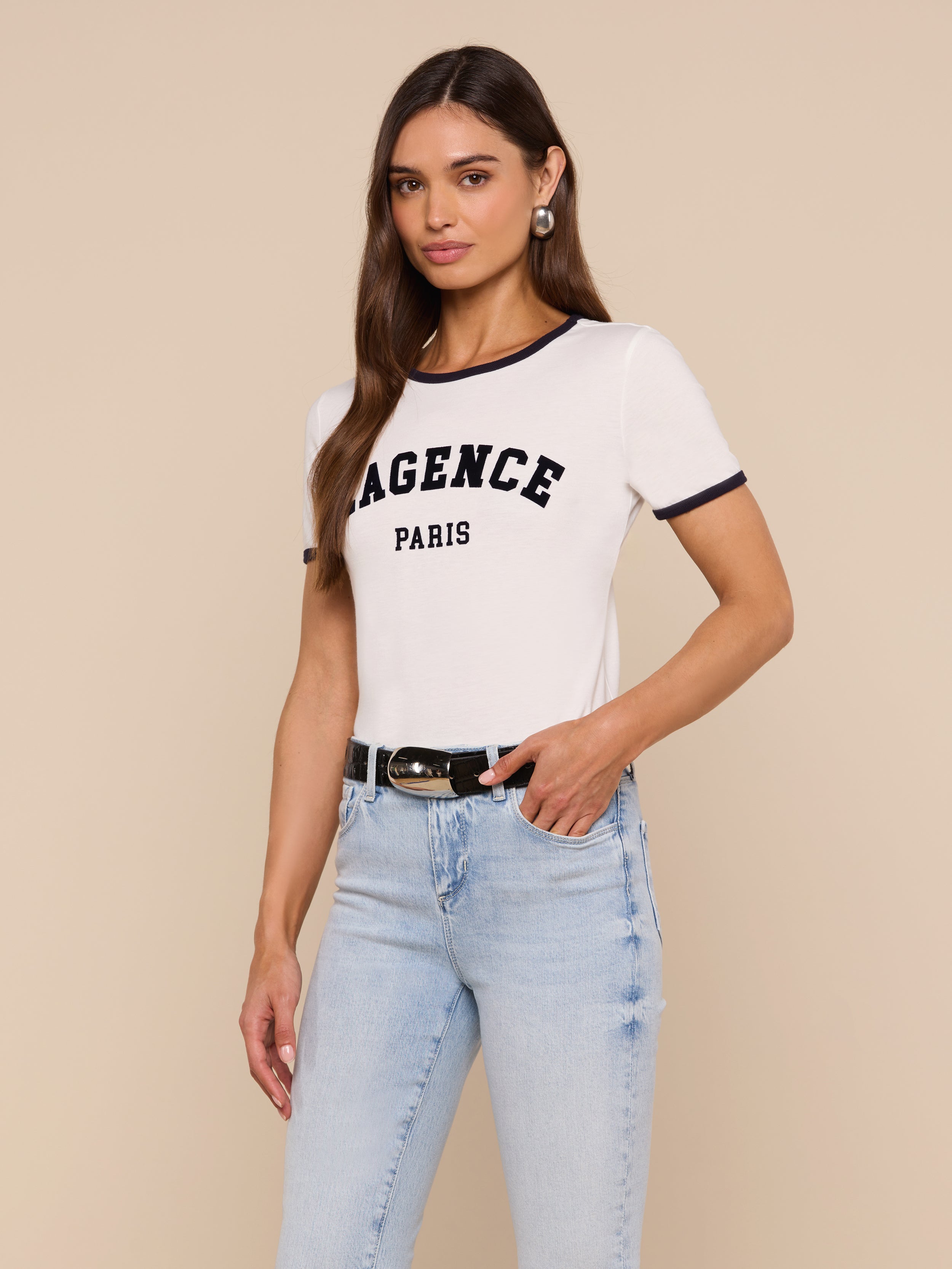 Woman wearing the Tommie Crewneck Tee in white with LAGENCE PARIS text, light blue jeans, black belt, and hoop earrings, standing against a plain light background in a relaxed fit Supima cotton style.