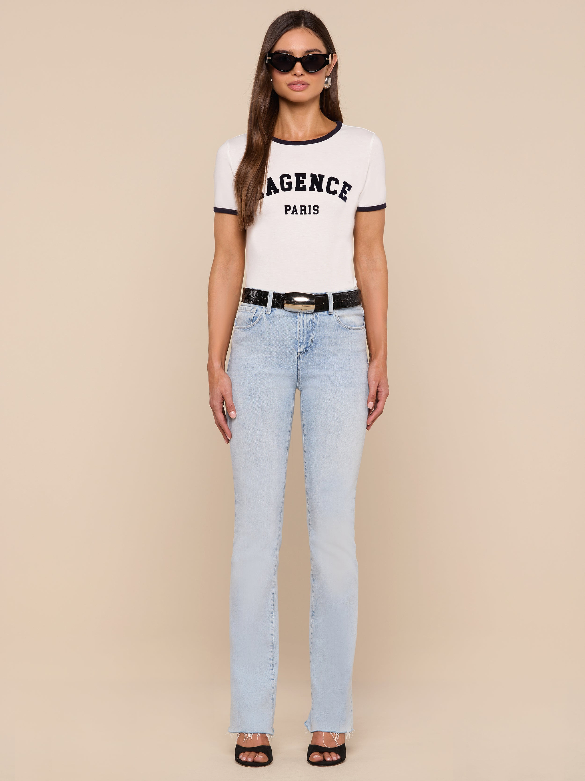 A woman stands against a plain white background wearing sunglasses, light blue jeans with a black belt, black shoes, and the Tommie Crewneck Tee—a relaxed fit Paris tee made from Supima cotton featuring the AGENCE PARIS text.