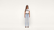 A woman stands against a plain white background, wearing sunglasses, a white AGENCY PARIS T-shirt, light blue jeans, a black belt, and heeled sandals.