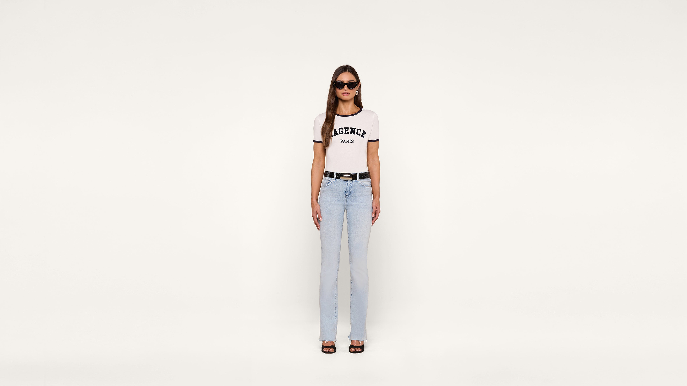 A woman stands against a plain white background, wearing sunglasses, a white AGENCY PARIS T-shirt, light blue jeans, a black belt, and heeled sandals.