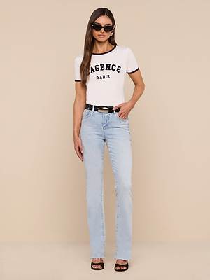 A woman stands against a beige background wearing black sunglasses, a relaxed-fit Tommie Crewneck Tee with L’AGENCE PARIS in black letters, light blue high-waisted jeans, a black belt, and black heeled sandals.