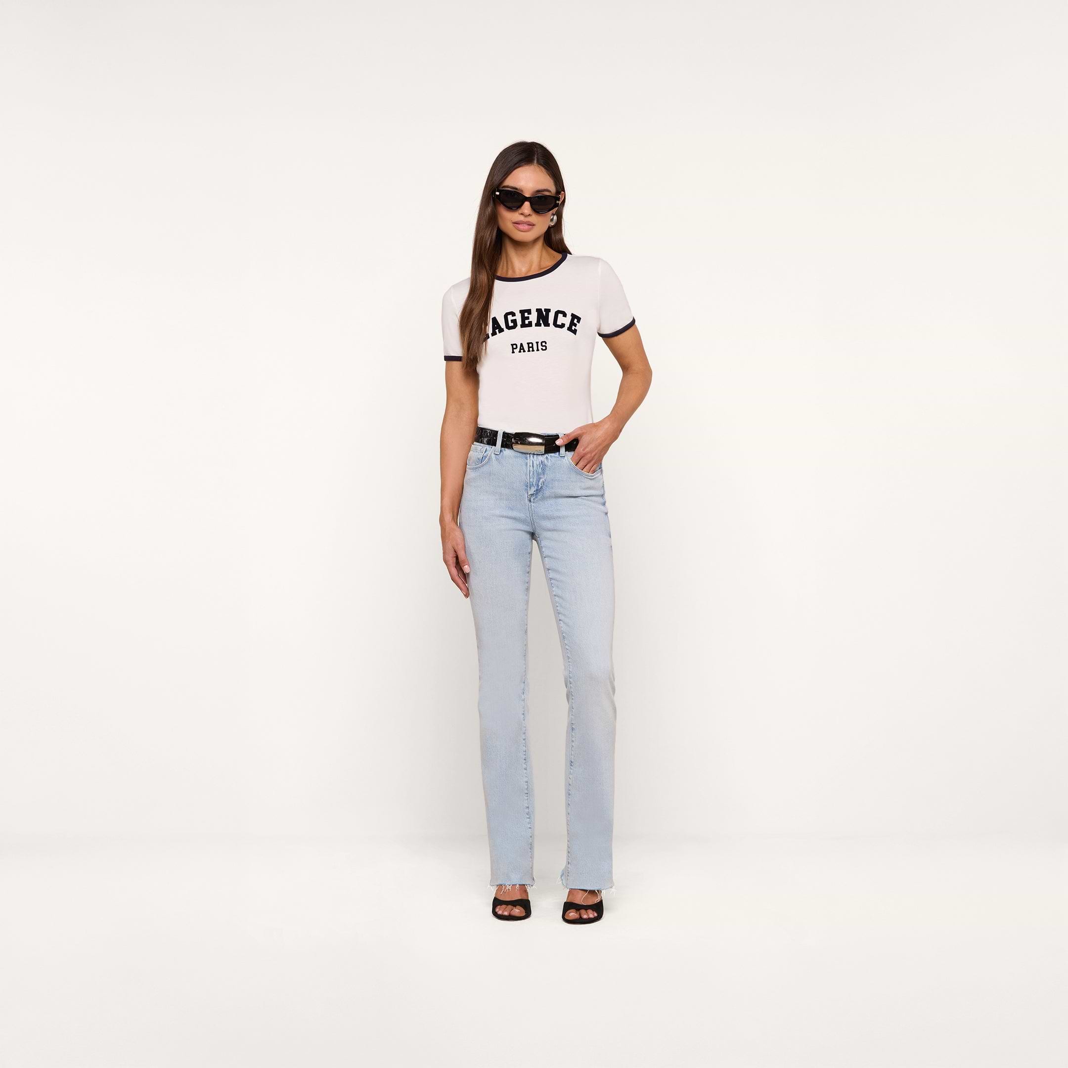 A woman stands against a plain white background wearing sunglasses, light blue jeans with a black belt, black sandals, and the white Tommie Crewneck Tee made from Supima cotton.