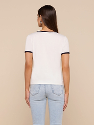 A woman with long brown hair, wearing a relaxed fit Tommie Crewneck Tee in white with black trim and light blue jeans, stands facing away from the camera against a plain background.