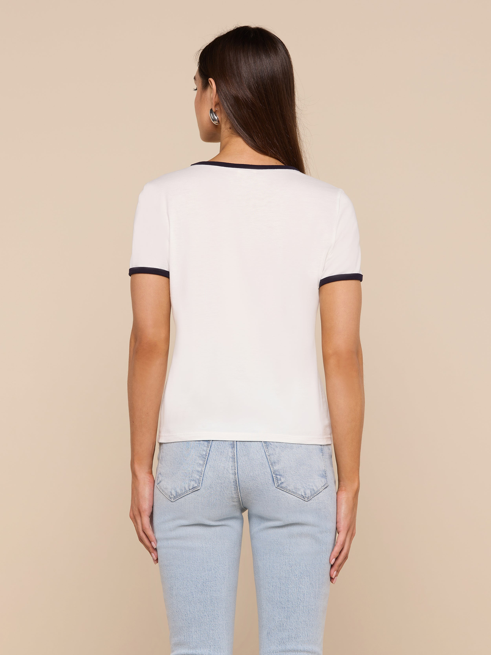 A woman with long brown hair, wearing a relaxed fit Tommie Crewneck Tee in white with black trim and light blue jeans, stands facing away from the camera against a plain background.