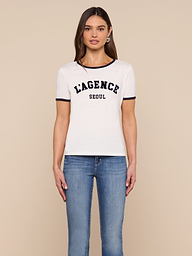 A woman stands against a plain background, wearing the Tommie Crewneck Tee—a relaxed fit white ringer tee with black trim that reads “LAGENCE SEOUL”—paired with blue jeans, looking forward with her arms relaxed by her sides.