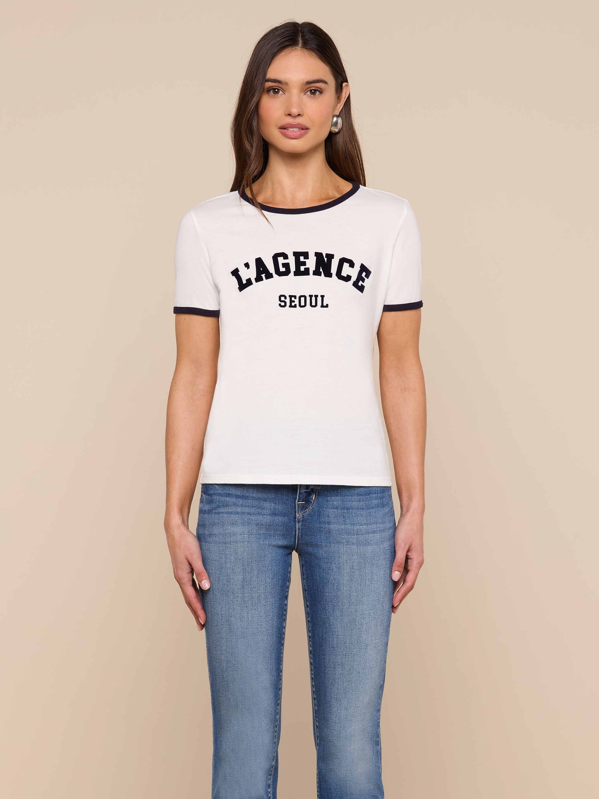 A woman stands against a plain background, wearing the Tommie Crewneck Tee—a relaxed fit white ringer tee with black trim that reads “LAGENCE SEOUL”—paired with blue jeans, looking forward with her arms relaxed by her sides.