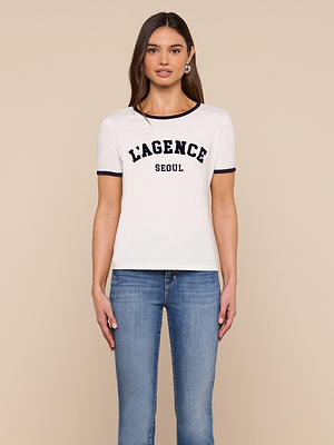 A woman with long brown hair wears the Tommie Crewneck Tee, a relaxed-fit L’AGENCE top in soft Supima cotton with black trim and LAGENCE SEOUL on the front, paired with blue jeans against a beige background.