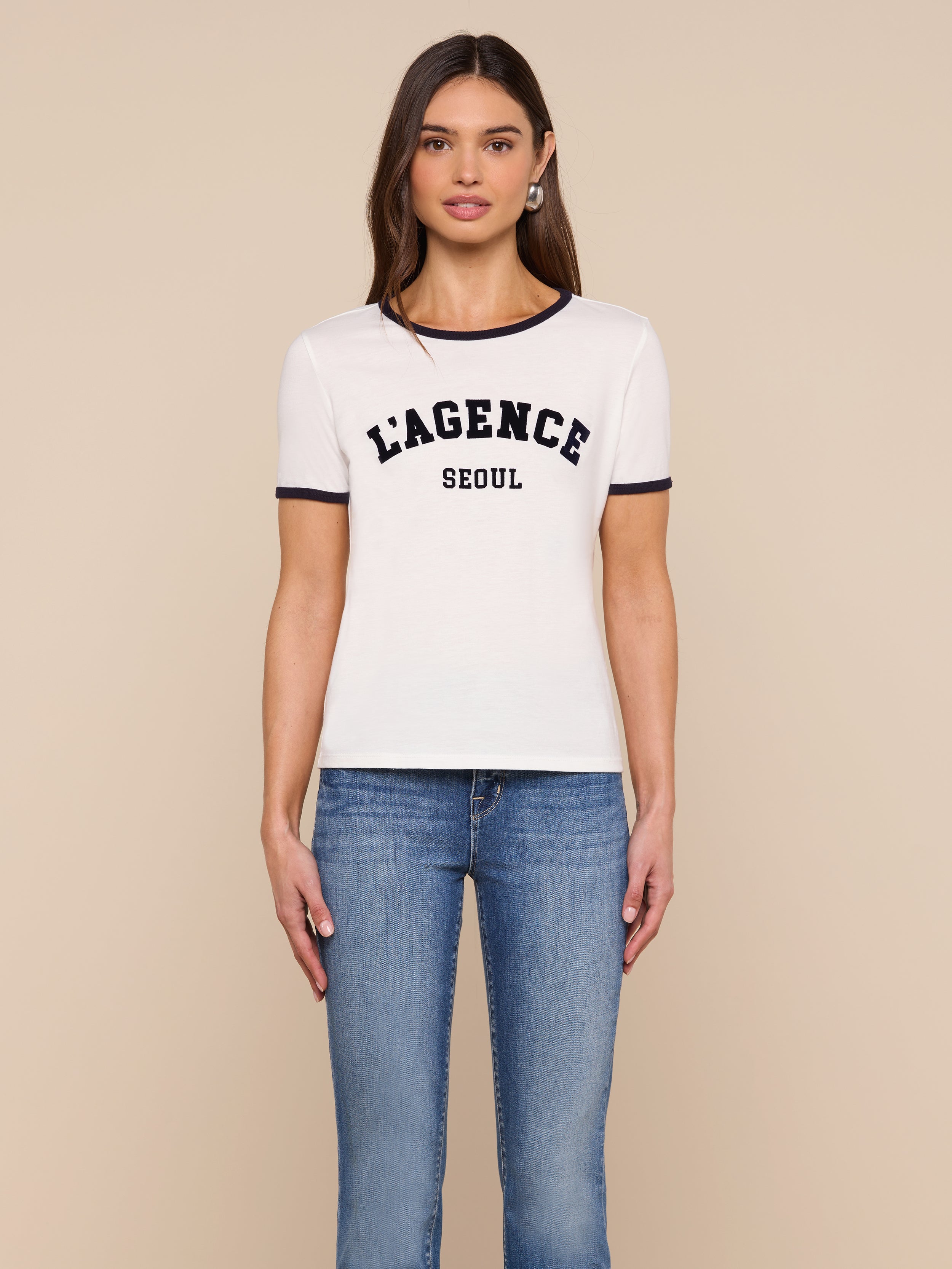 A woman stands against a plain background, wearing the Tommie Crewneck Tee—a relaxed fit white ringer tee with black trim that reads “LAGENCE SEOUL”—paired with blue jeans, looking forward with her arms relaxed by her sides.