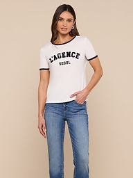A woman stands against a plain background, wearing the Tommie Crewneck Tee—a white ringer style with “LAGENCE SEOUL” printed on the front—paired with blue jeans. She has one hand in her pocket and a relaxed expression.