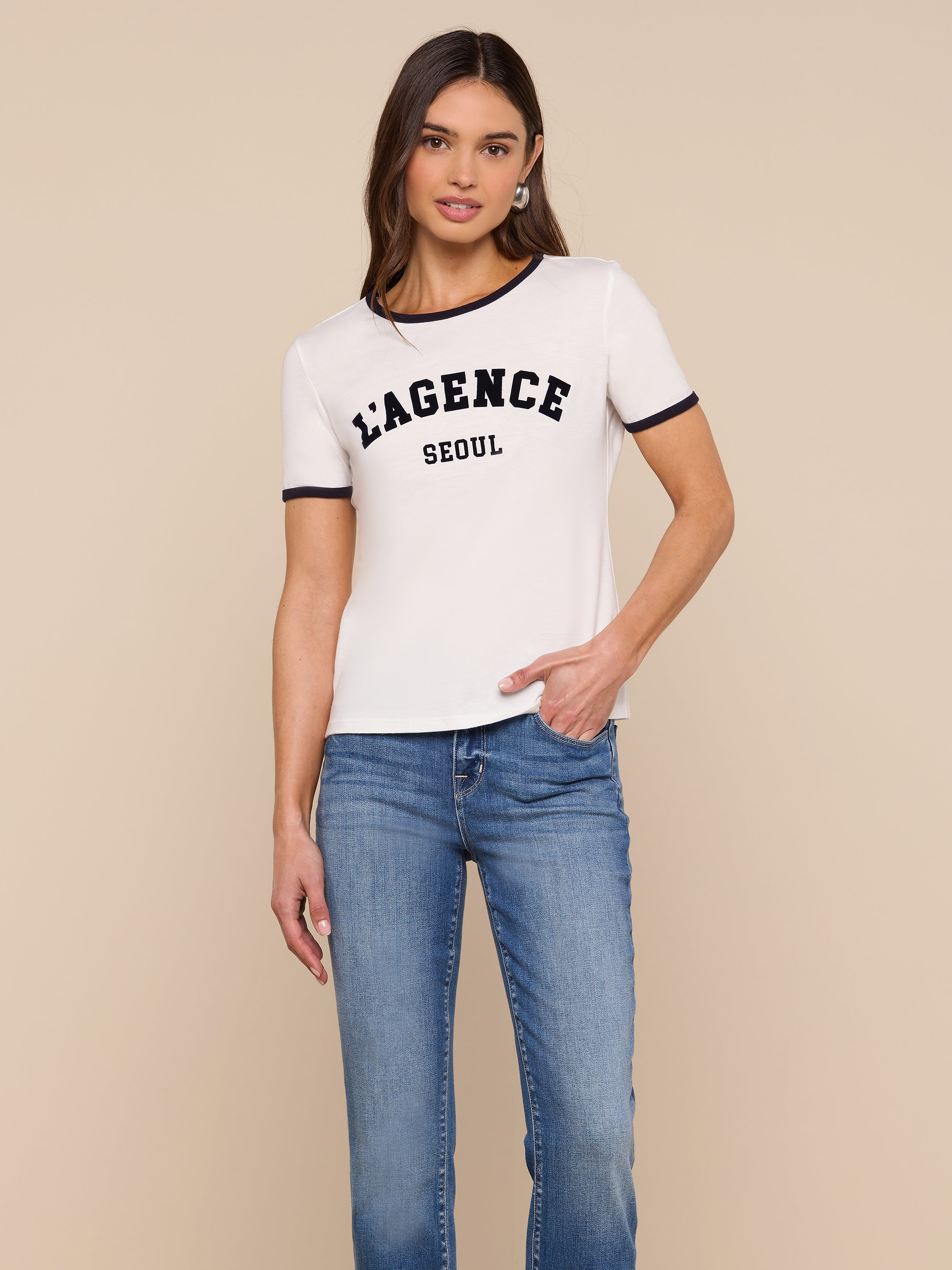 A woman stands against a plain background, wearing the Tommie Crewneck Tee—a white ringer style with “LAGENCE SEOUL” printed on the front—paired with blue jeans. She has one hand in her pocket and a relaxed expression.