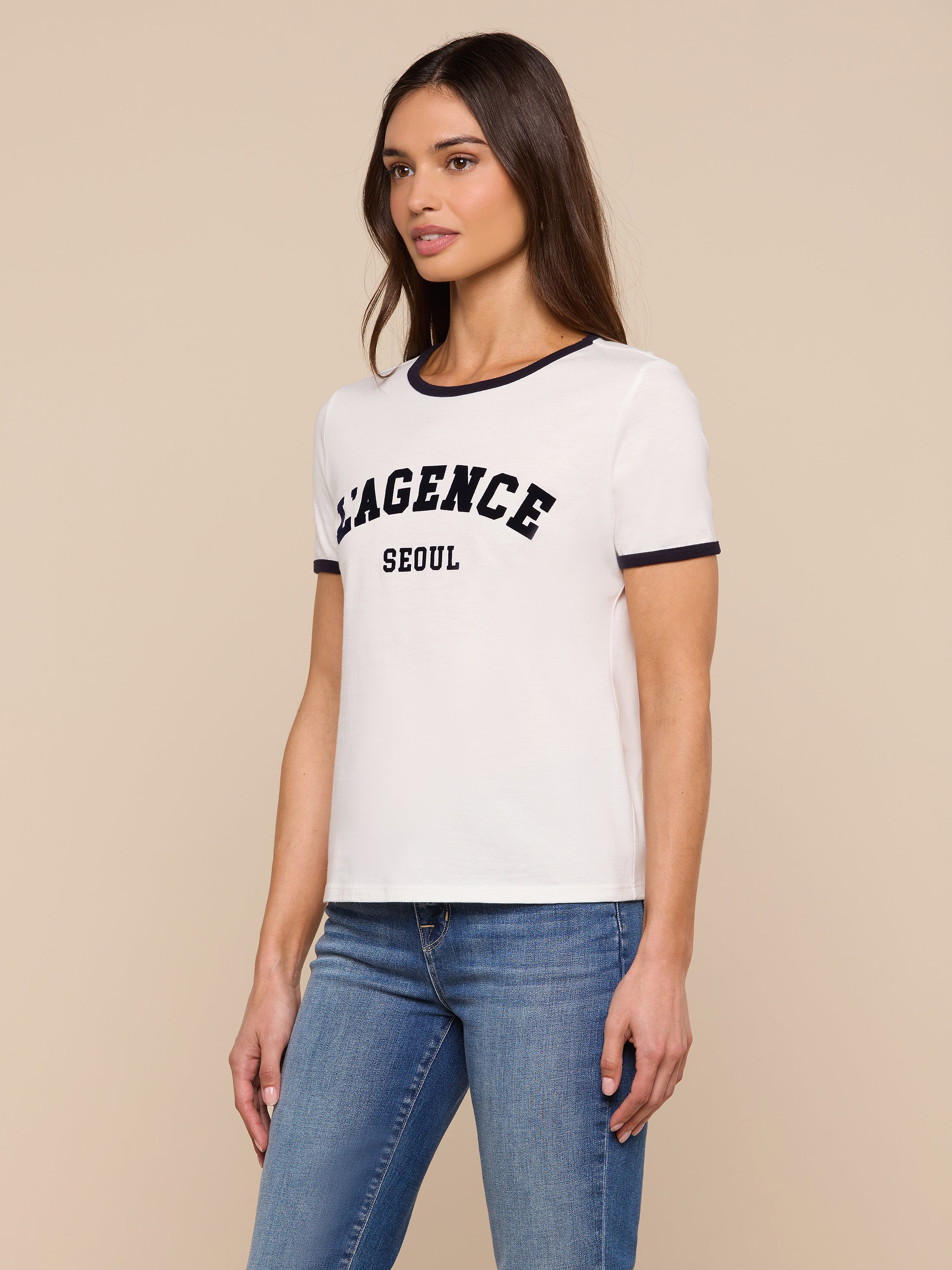 A woman wearing a white Tommie Crewneck Tee made from Supima cotton, featuring “LAGENCE SEOUL” printed on the front, and blue jeans stands against a plain background, gazing slightly to her right. The relaxed fit of the tee enhances her casual, effortless style.