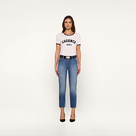 A woman stands facing forward, wearing a white Tommie Crewneck Tee in Supima cotton with a relaxed fit, blue jeans, a black belt, and black high heels against a plain white background.