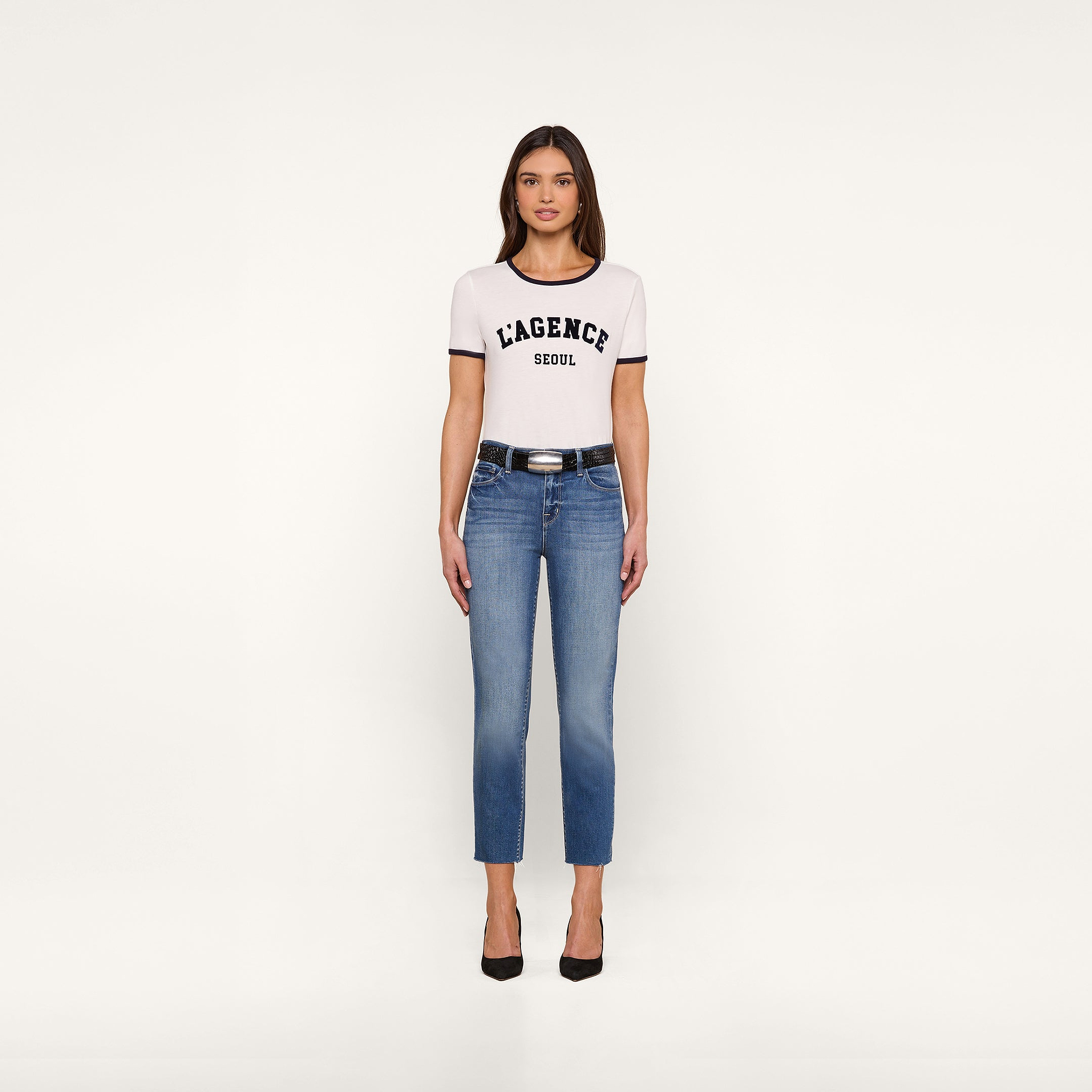 A woman stands facing forward, wearing a white Tommie Crewneck Tee in Supima cotton with a relaxed fit, blue jeans, a black belt, and black high heels against a plain white background.