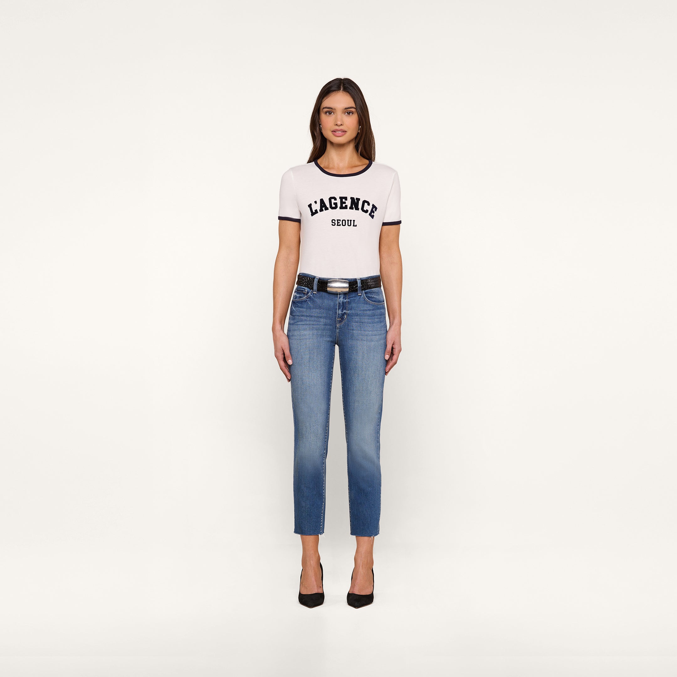 A woman stands facing forward, wearing a white Tommie Crewneck Tee in Supima cotton with a relaxed fit, blue jeans, a black belt, and black high heels against a plain white background.