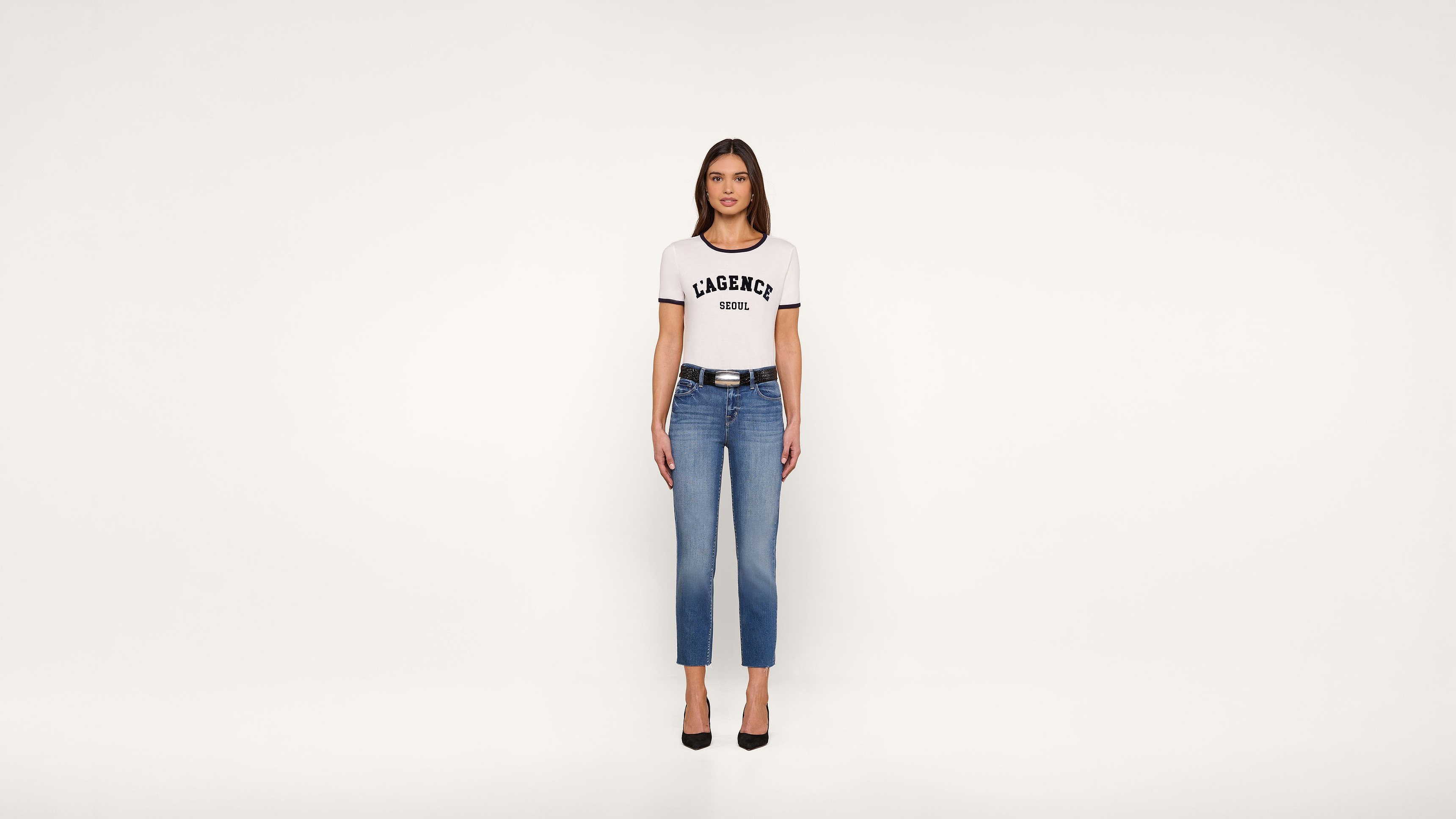 A woman stands facing forward, wearing a white t-shirt with LACERNE SCHOOL text, blue jeans, a black belt, and black heels, against a plain white background.