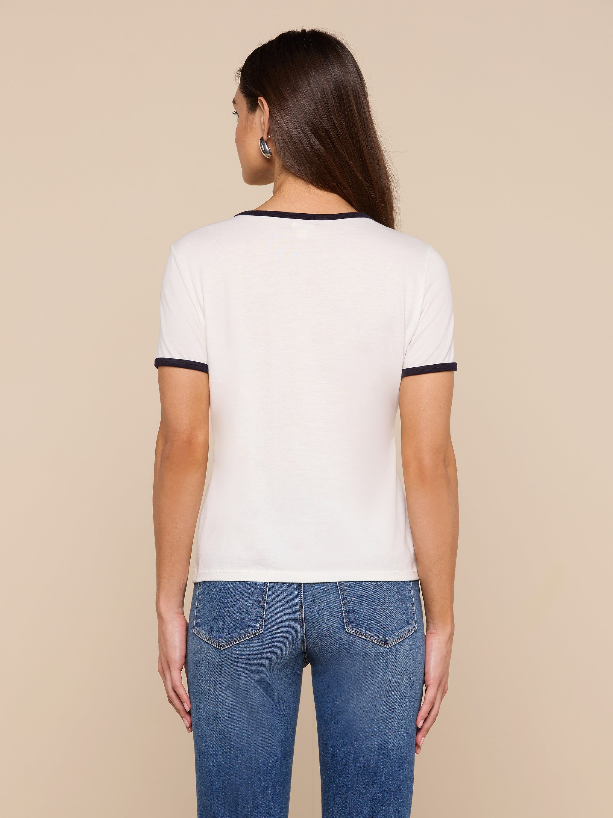 A woman with long brown hair stands facing away, wearing the Tommie Crewneck Tee—crafted from soft Supima cotton in a relaxed fit—paired with blue jeans against a plain light background.