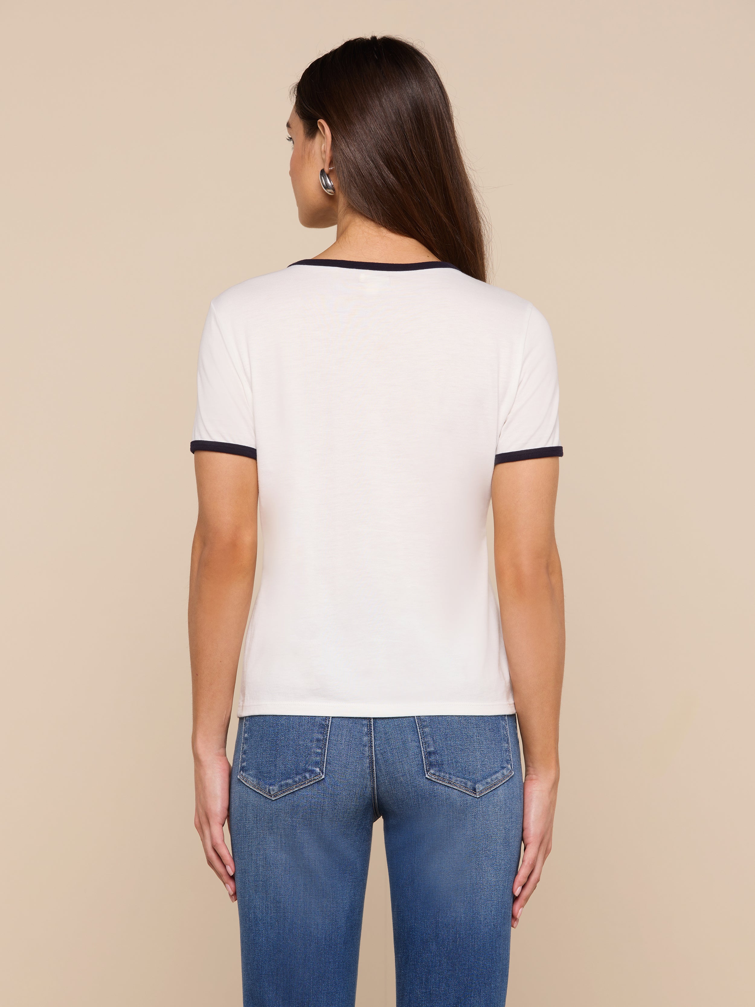 A woman with long brown hair stands facing away, wearing the Tommie Crewneck Tee—crafted from soft Supima cotton in a relaxed fit—paired with blue jeans against a plain light background.