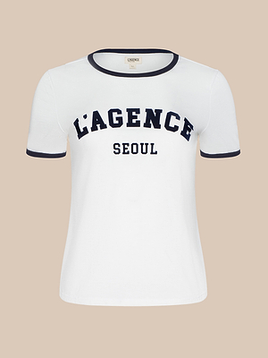 The Tommie Crewneck Tee is a relaxed fit white L’AGENCE tee in soft Supima cotton, with black trim on the collar and sleeves. LAGENCE appears in large black letters and SEOUL in smaller black letters on the front.