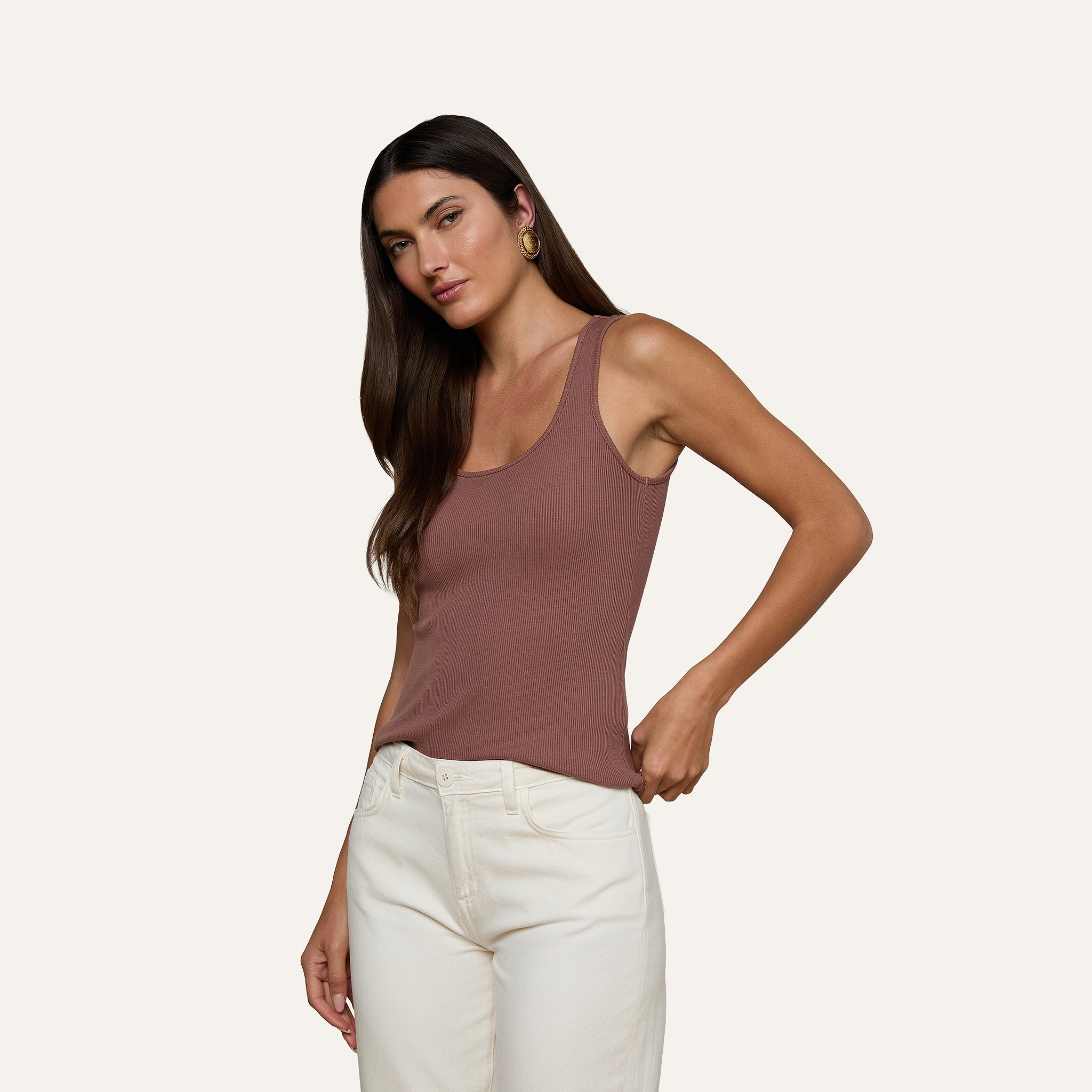 Una donna dai lunghi capelli scuri indossa la canotta Adira Scoopneck Tank in un intramontabile color malva, abbinata a pantaloni bianchi, e posa su uno sfondo semplice guardando leggermente di lato con una mano in tasca. Il tessuto a costine elasticizzato della canotta Adira Scoopneck Tank aggiunge comfort e stile al suo look disinvolto.