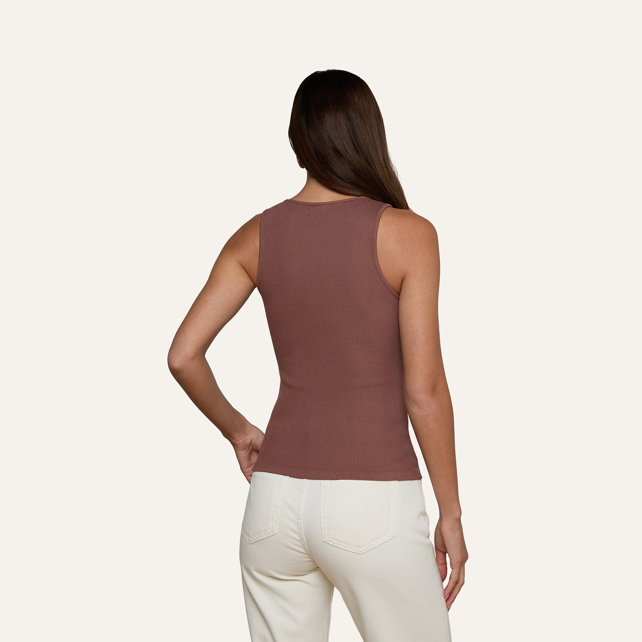 Una donna dai lunghi capelli castani, che indossa la canotta Adira Scoopneck e pantaloni bianchi, è in piedi di spalle alla fotocamera su uno sfondo chiaro e uniforme.